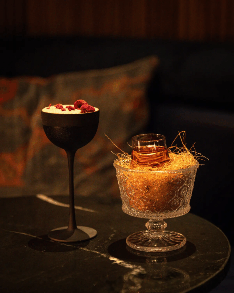 Two artisanal cocktails on a dark marble table, one garnished with berries in a black glass and the other in a crystal glass with straw nest
