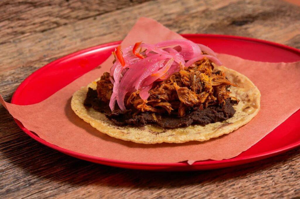 Close-up of a traditional taco with black beans, shredded meat, and pickled red onions on a red plate