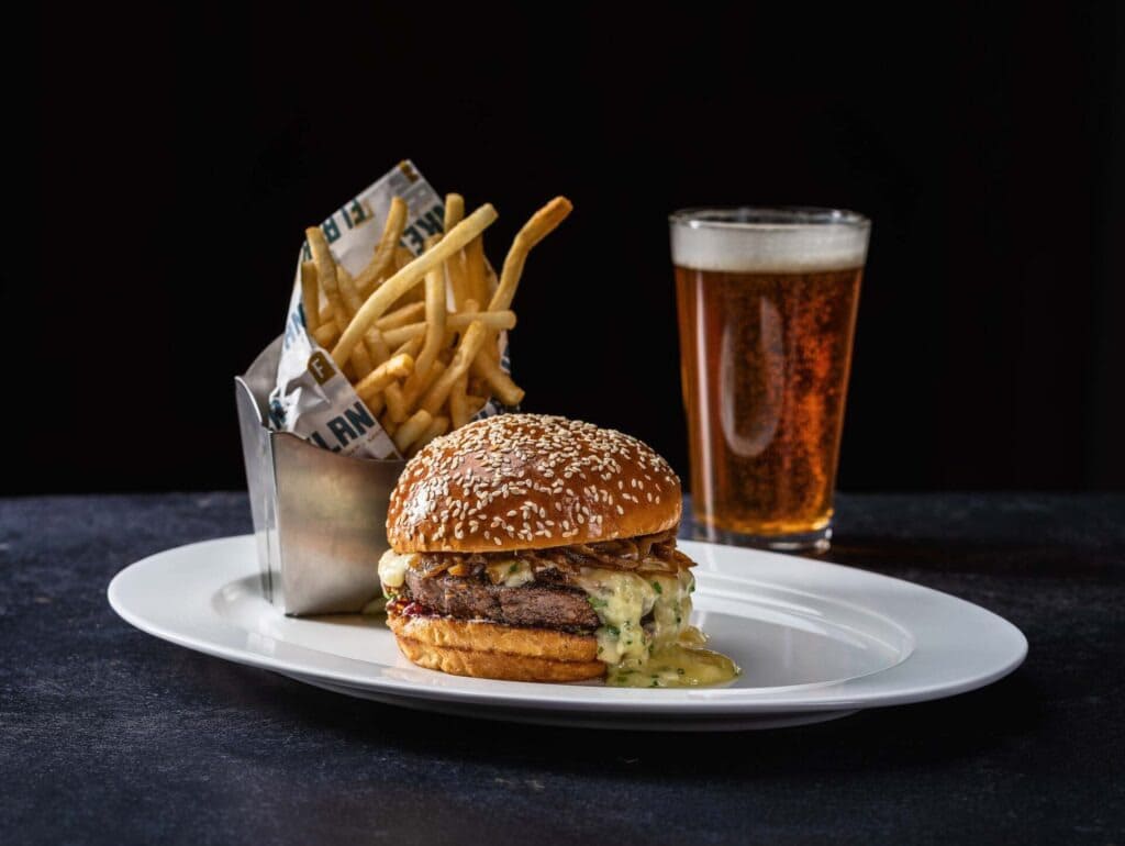 Gourmet burger with cheese and sauce on a sesame bun, served with fries in a Flanker wrapper and a glass of beer on a white plate.