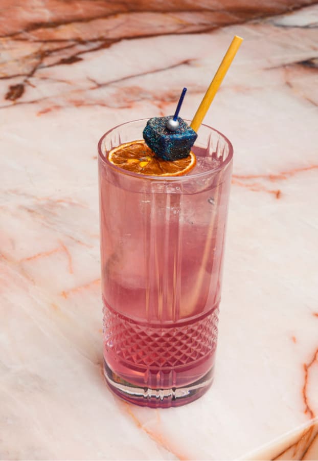Pink cocktail in a textured glass with a dried orange slice and garnish on a marble surface