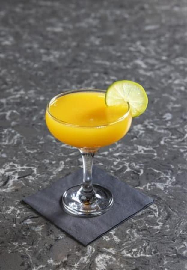 Yellow citrus cocktail in a coupe glass garnished with a lime wheel on a dark stone surface.