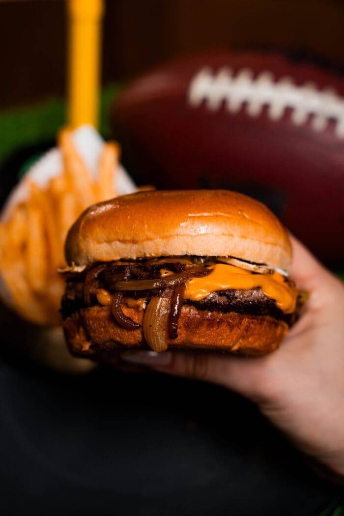 Close-up of a cheeseburger with grilled onions held in front of a football and fries.