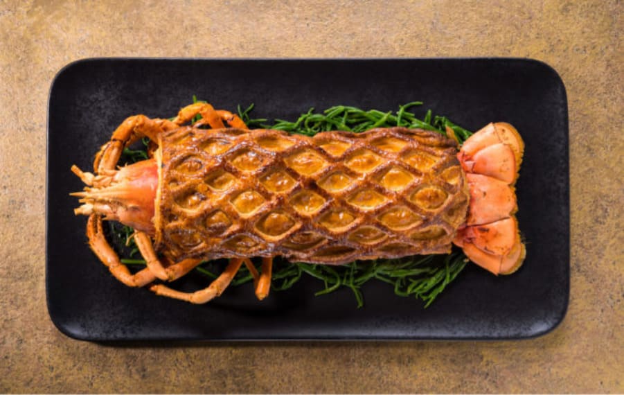 Whole grilled lobster arranged on a black rectangular plate over a bed of green herbs.