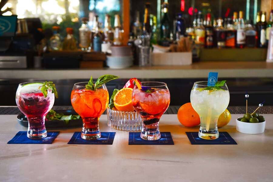 Four cocktails with unique garnishes lined up on a bar, with fruits, herbs, and snacks in the background.
