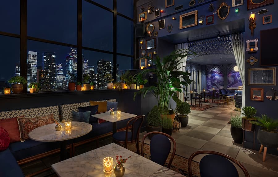 Elegant lounge interior with marble tables, candle lighting, indoor plants, and a nighttime city skyline view through large windows.