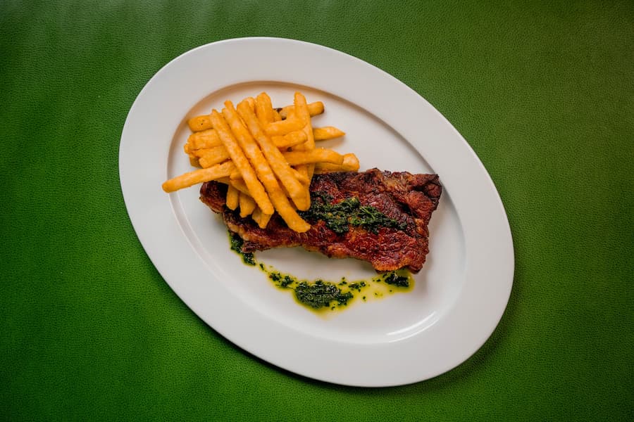 Grilled steak topped with herb sauce served with French fries on a white plate.