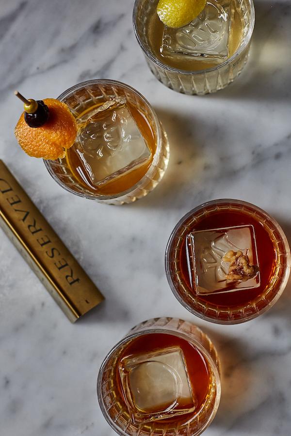 Overhead view of four handcrafted cocktails served over large ice cubes on a marble bar top, garnished with citrus peel, cherry, and walnut accents.