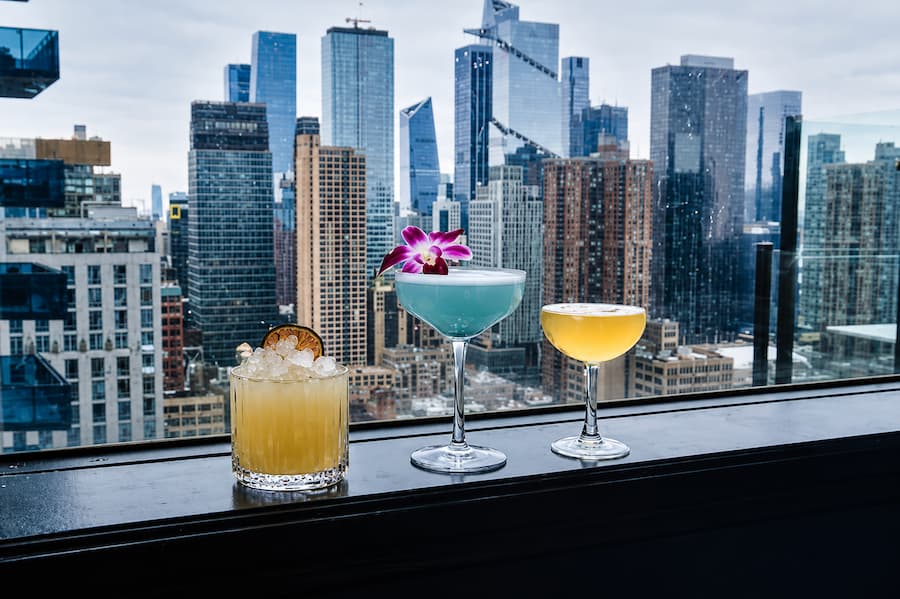 Three handcrafted cocktails displayed on a rooftop ledge overlooking a modern city skyline with high-rise buildings in the background.