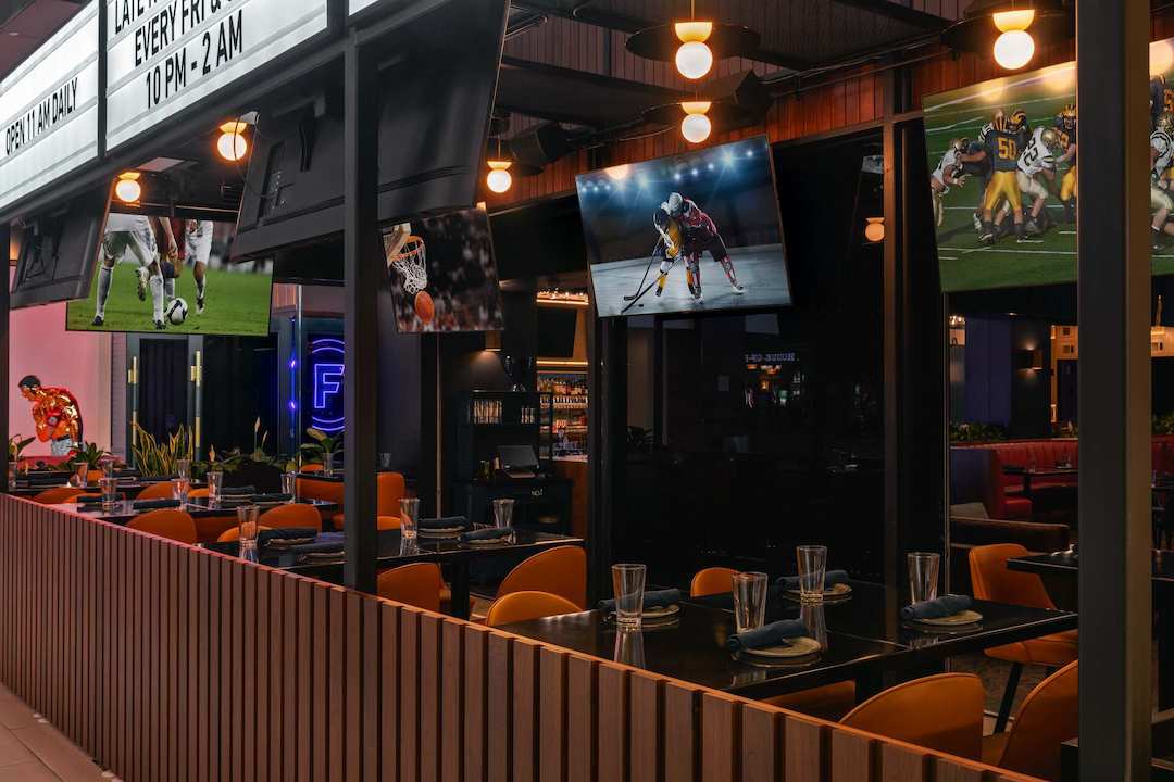 Modern sports bar dining area with tables, orange chairs, and multiple large televisions displaying live sporting events, creating a lively atmosphere for watching games while dining.