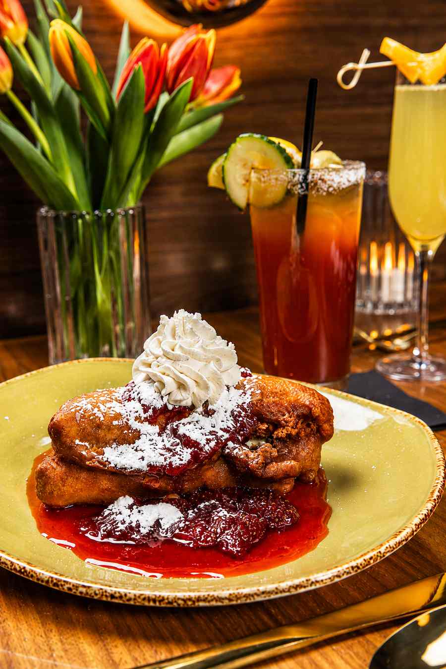 French toast topped with berry sauce, powdered sugar, and whipped cream, served alongside colorful brunch cocktails and fresh flowers.