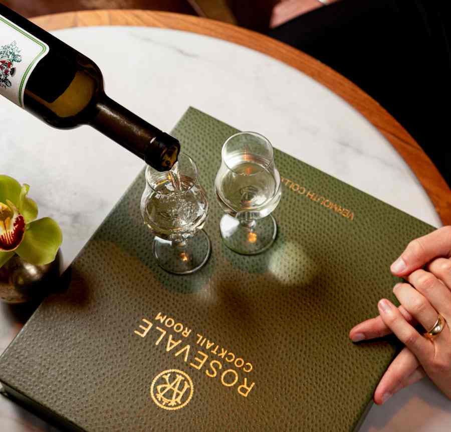 Wine being poured into a glass beside a Rosevale Cocktail Room menu on a marble table.