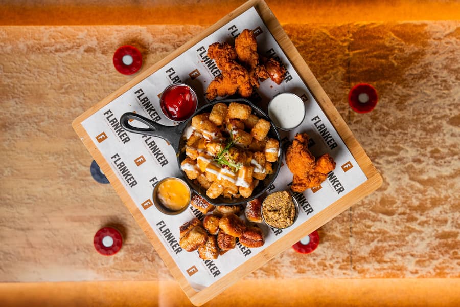 Overhead view of burgers, fries, wings, chicken tenders, steak, pretzel, drinks, and sports-themed decor arranged on a table.